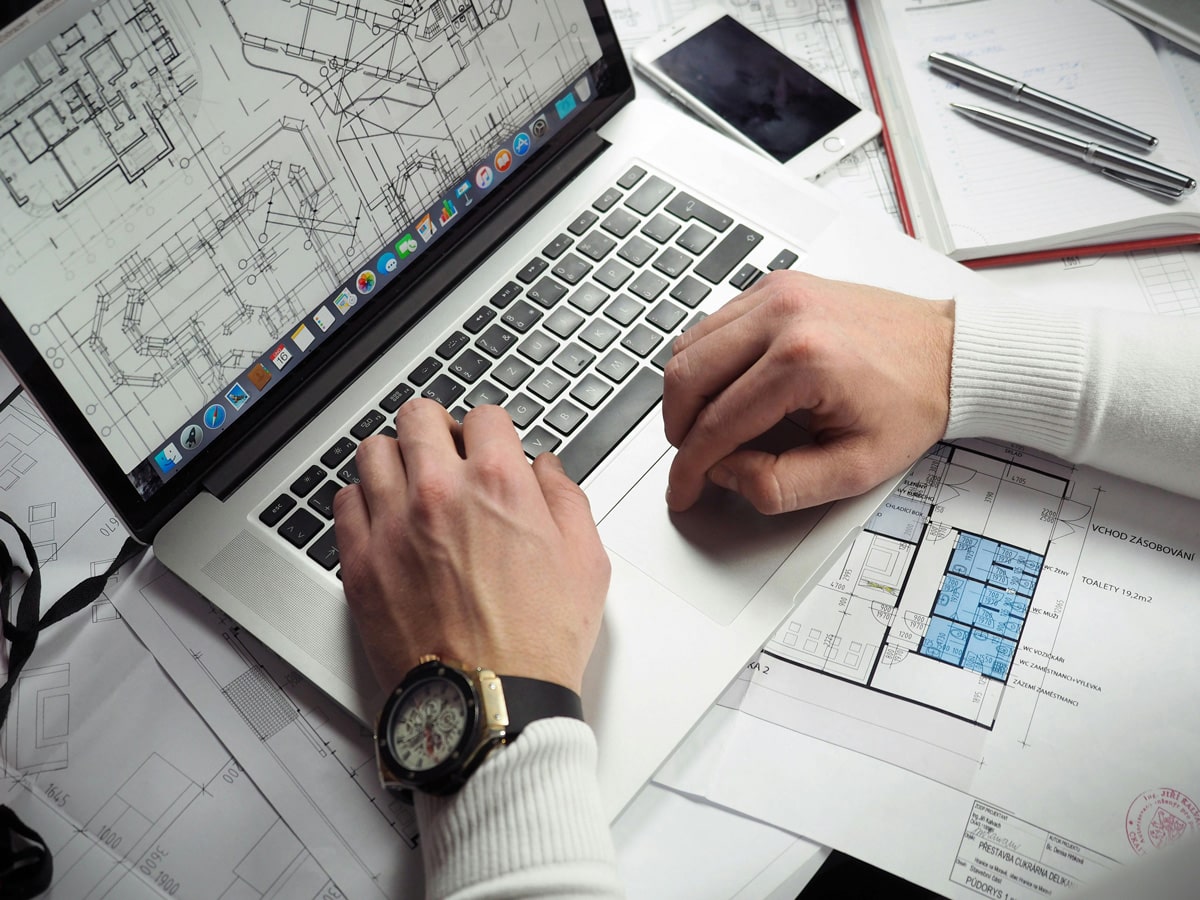 man working on construction drawings on laptop computer with print outs of drawings underneath computer on desktop with drafting tools